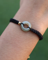 A close-up of a person's wrist wearing a black braided cord bracelet with a circular light gray stone centerpiece and a small gold accent.